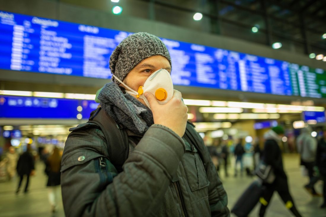 Man wearing a respirator mask device for health protection at an airport or railway train station in a crowd of people while travelling. Epidemic corona virus infection, flu sickness and travel illness concept.